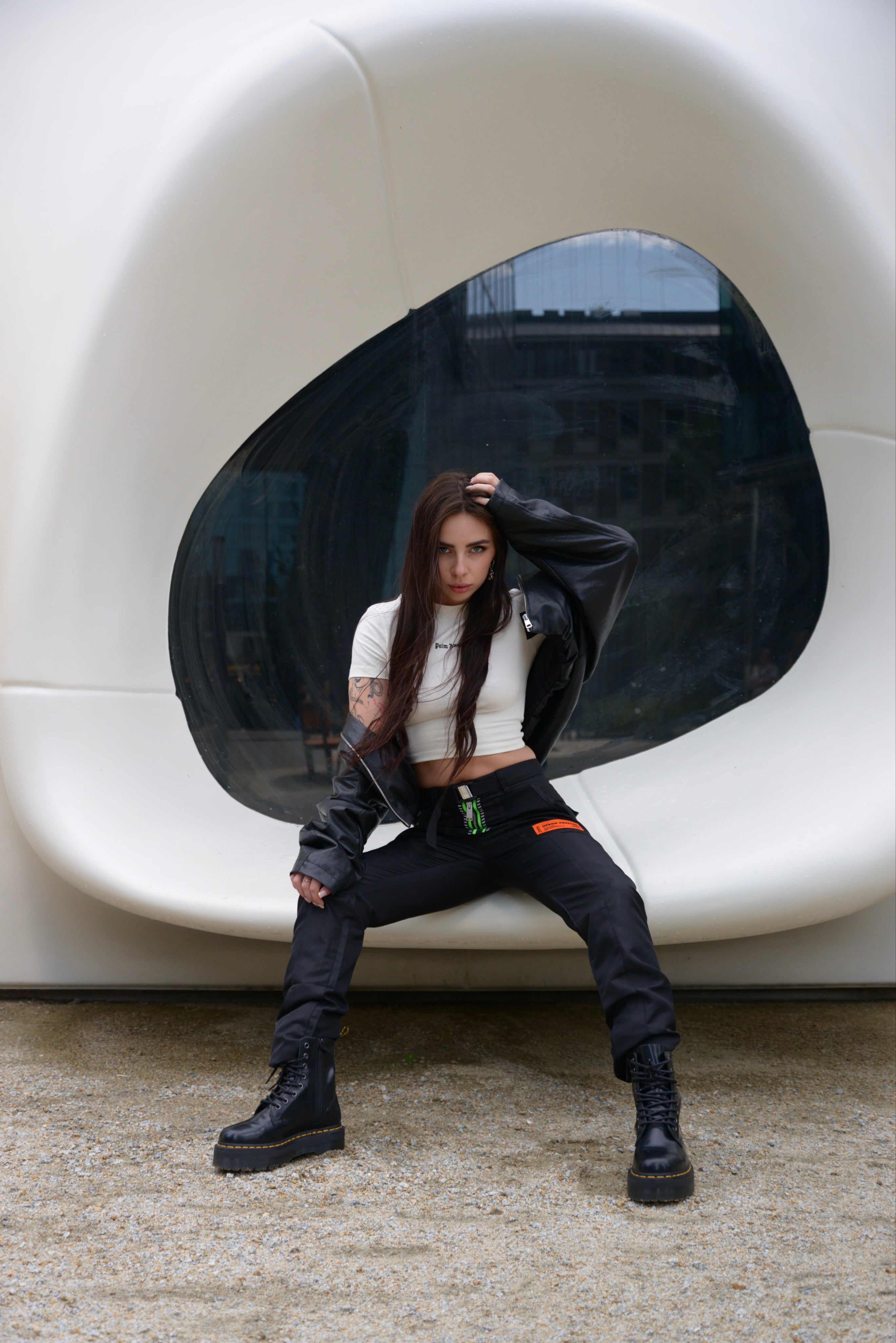 Lizaveta Kaval in a black leather jacket and cargo trousers, seated in front of a white sculptural pavilion