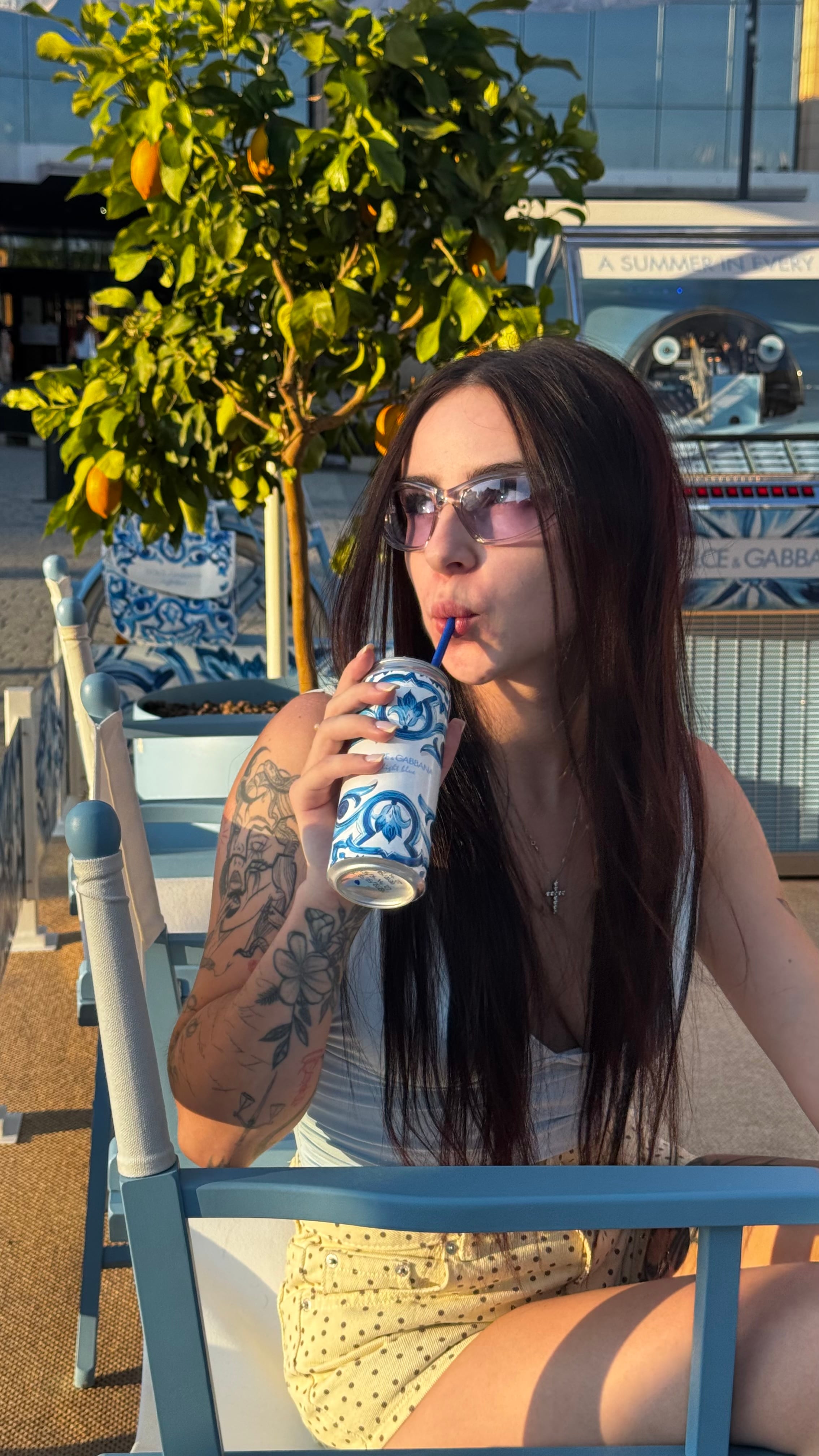 Lizaveta in tinted sunglasses drinking from a blue-and-white printed can on a sunny terrace