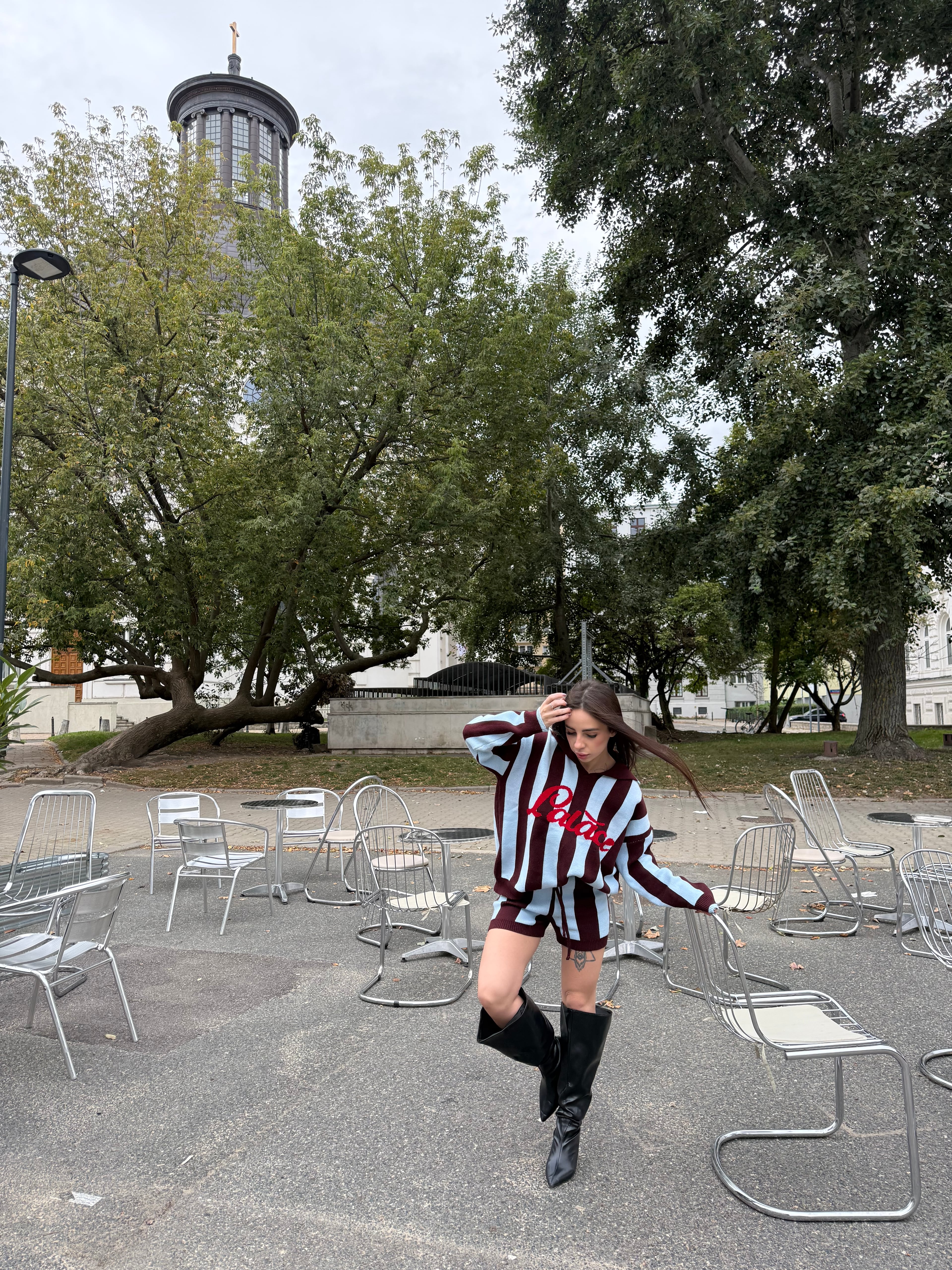 Lizaveta in a striped burgundy knit set and knee-high black boots among metal chairs in a park