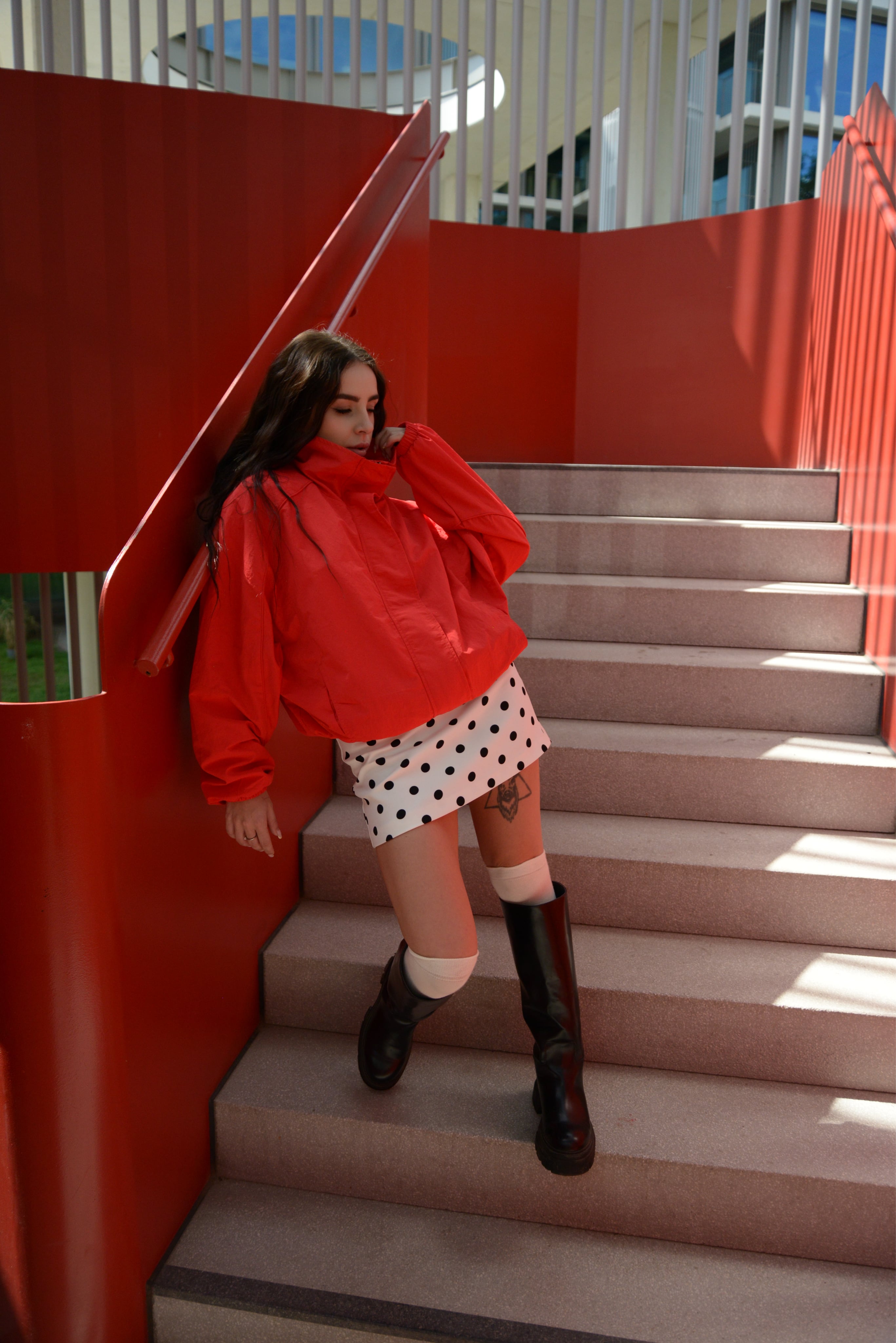Lizaveta in a red jacket and polka-dot skirt leaning on a red staircase