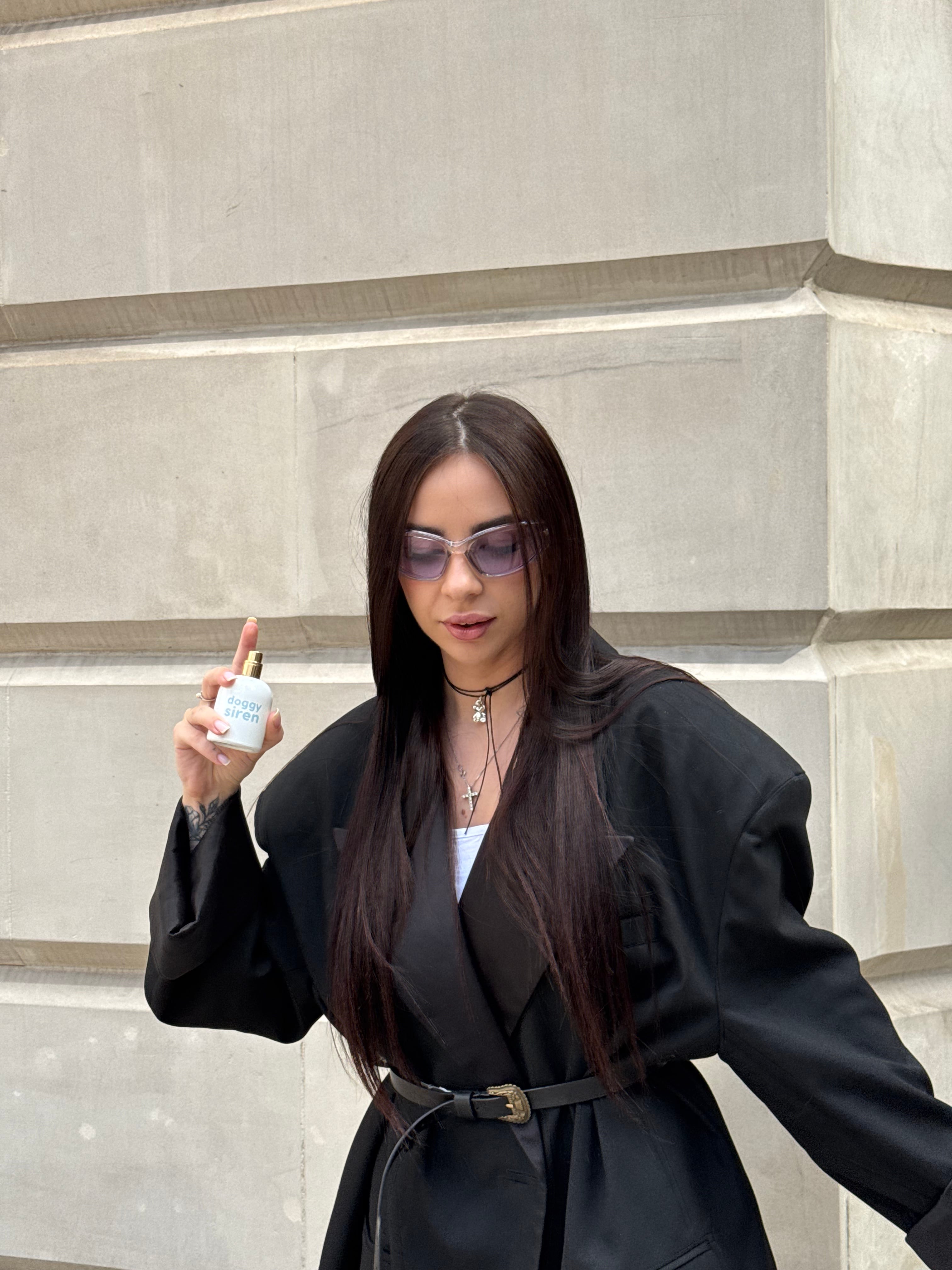 Lizaveta in an oversized black blazer and tinted glasses holding up a perfume bottle